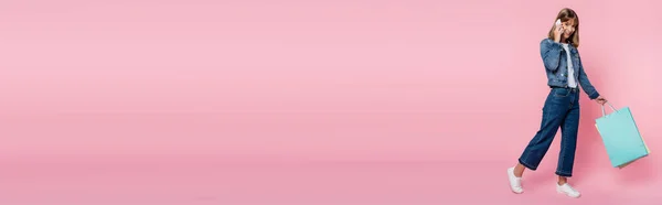 Colpo panoramico di donna in giacca di denim che parla su smartphone e tiene borse della spesa su sfondo rosa — Foto stock