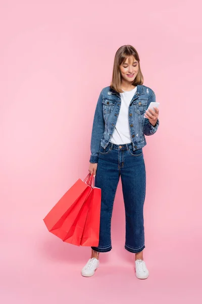 Frau in Jeans und Jeansjacke nutzt Smartphone und hält rote Einkaufstüten auf rosa Hintergrund — Stockfoto