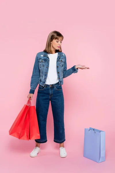 Mujer joven sosteniendo bolsas de compras y señalando con la mano sobre fondo rosa - foto de stock