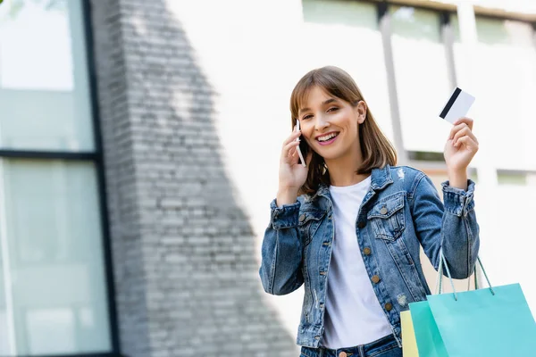 Junge Frau zeigt Kreditkarte, während sie Einkaufstüten hält und auf der Straße mit dem Smartphone spricht — Stockfoto