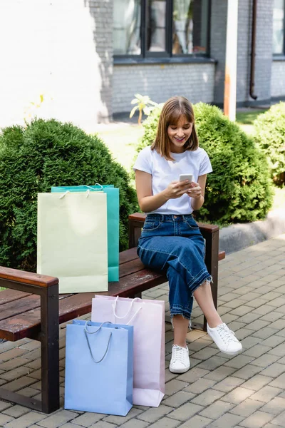 Young woman using smartphone near shopping bags on bench on urban street — Stock Photo