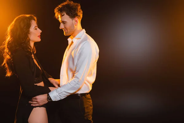 Seductive woman in blazer looking at boyfriend in shirt on black — Stock Photo