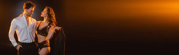 Панорамний знімок кучерявої жінки в мереживному нижній білизні, що тримає світло, дивлячись на чоловіка, що стоїть з рукою в кишені на чорному — стокове фото