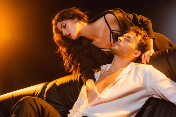 Curly woman in lace bra and blazer touching man in shirt sitting on sofa on black — Stock Photo