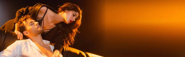 Panoramic crop of curly woman in lace bra and blazer touching man in shirt sitting on sofa on black — Stock Photo