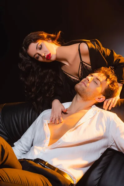 Seductive woman in lace bra and blazer touching man with closed eyes sitting on sofa on black — Stock Photo