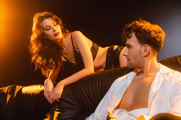 Man in shirt sitting on sofa and looking at seductive woman in lace underwear on black — Stock Photo