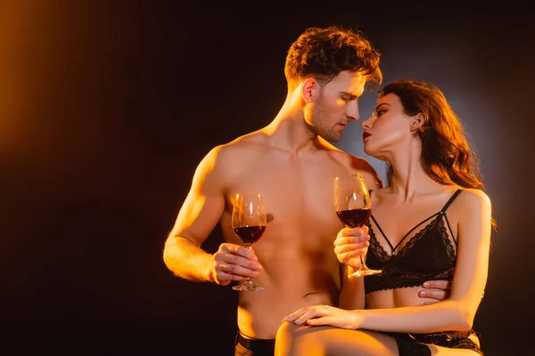 Мужчина без рубашки и соблазнительная женщина, держащие бокалы с красным вином, глядя друг на друга на черном — стоковое фото