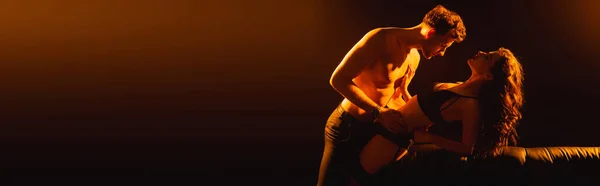 Panoramisches Konzept: muskulöser Mann umarmt sexy Frau in Unterwäsche in der Nähe von Sofa auf schwarz — Stockfoto