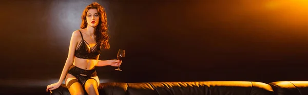 Panoramic shot of seductive woman in underwear sitting on sofa and holding glass of red wine on black — Stock Photo