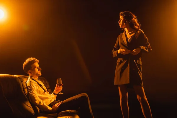 Curly woman touching jacket and looking at man with glass of red wine sitting on black — Stock Photo