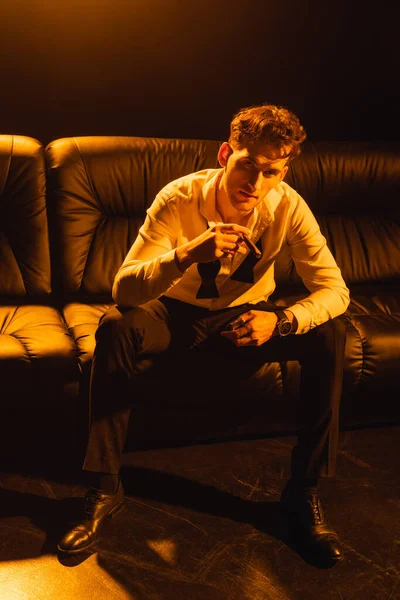 Man in formal wear holding cigar, sitting on sofa and looking at camera on black — Stock Photo