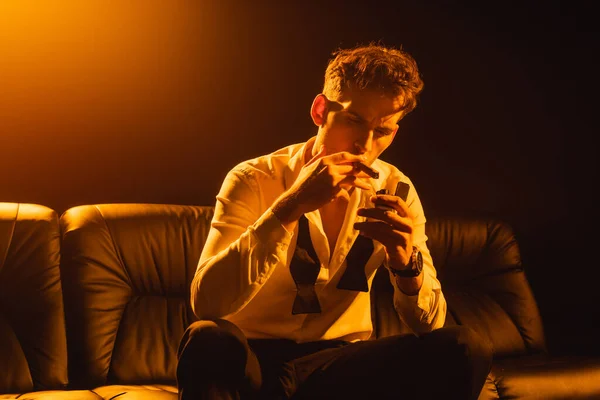 Man in formal wear holding lighter near cigar while sitting on sofa on black — Stock Photo