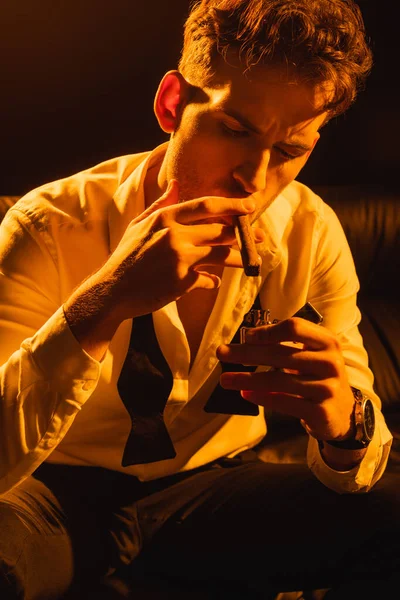 Hombre de traje sosteniendo encendedor cerca de cigarro mientras está sentado en el sofá en negro - foto de stock