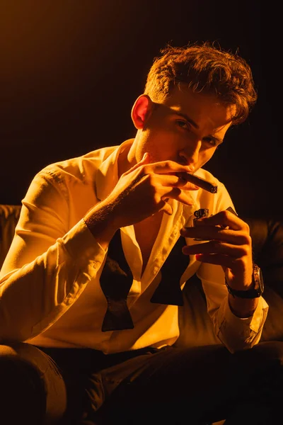 Hombre de traje sosteniendo más ligero cerca de cigarro mientras está sentado en el sofá y mirando a la cámara en negro - foto de stock