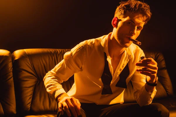 Man in suit holding lighter, smoking cigar, sitting on sofa and looking at camera on black — Stock Photo