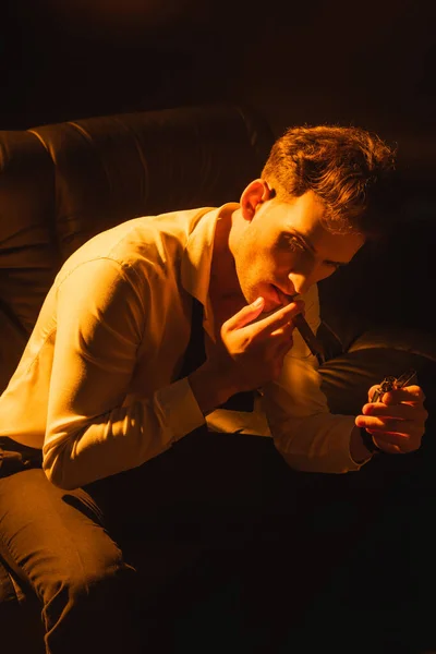 Mann im Anzug hält Feuerzeug in der Hand, raucht Zigarre und sitzt isoliert auf einem Sofa auf schwarz — Stockfoto