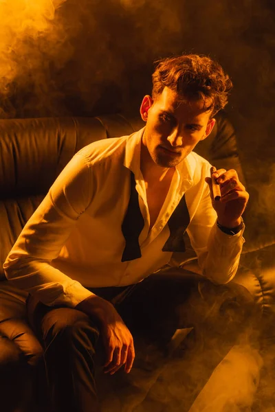 Man in suit sitting on sofa and holding cigar on black with smoke — Stock Photo