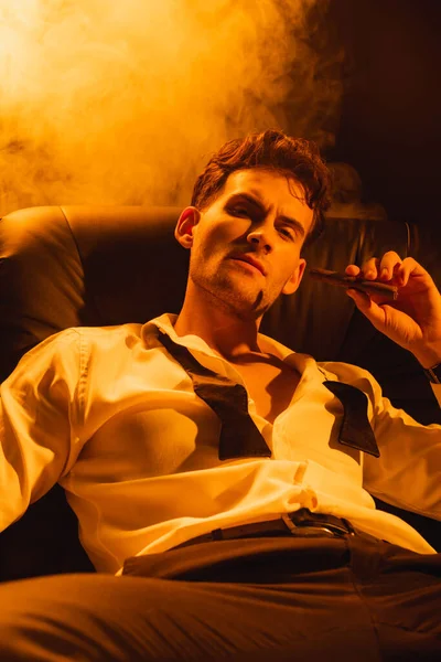 Hombre serio en traje sosteniendo cigarro y mirando a la cámara en negro con humo - foto de stock