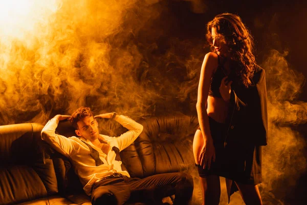 Man sitting on sofa and looking at seductive woman on black with smoke — Stock Photo