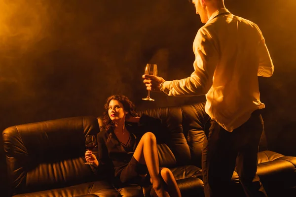 Hombre de traje sosteniendo copa de vino tinto cerca de mujer seductora en sujetador y chaqueta sentado en el sofá en negro con humo - foto de stock