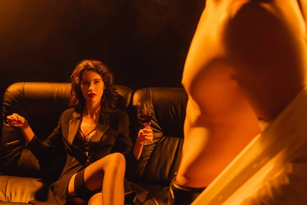 Selektiver Fokus der verführerischen Frau mit einem Glas Rotwein, die auf dem Sofa sitzt und muskulösen Mann ansieht, der sich auf schwarz auszieht — Stockfoto