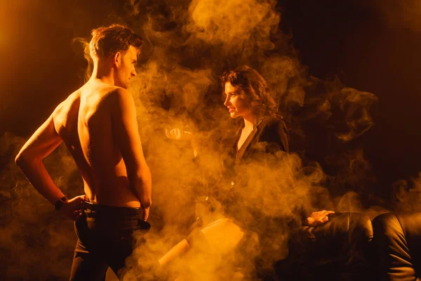 Lockige Frau in Jacke sieht einen Mann ohne Hemd an, der mit der Hand an der Hüfte auf schwarz mit Rauch steht — Stockfoto