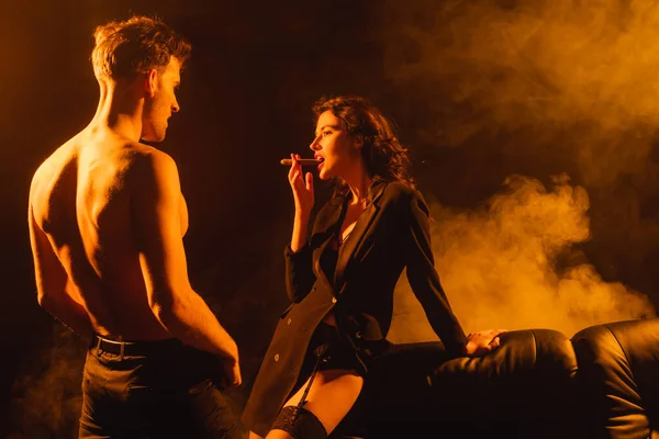 Seductora mujer fumando cigarro cerca de hombre sin camisa en negro — Stock Photo