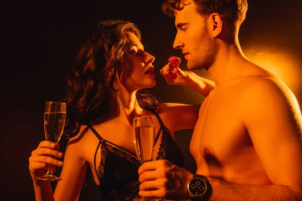 Спокуслива жінка тримає свіжу полуницю біля без сорочки чоловіка зі склянкою шампанського на чорному — стокове фото