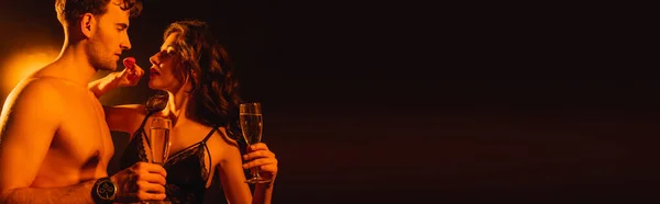 Horizontal crop of seductive woman holding fresh strawberry near shirtless man with glass of champagne on black — Stock Photo