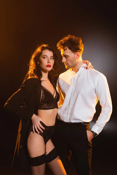 Man in suit standing with hand in pocket and looking at seductive woman in underwear and jacket on black — Stock Photo