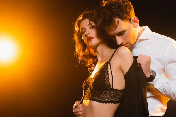 Passionate man in suit taking off jacket on sexy woman in bra on black with back light — Stock Photo