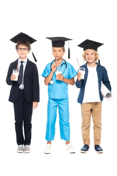 Crianças em gorros de graduação vestidos em trajes de profissões diferentes que mantêm copos com leite isolado no branco — Fotografia de Stock