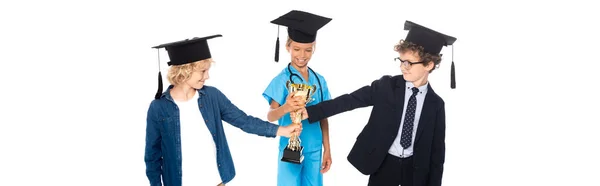 Панорамний знімок дітей у випускних кепках, одягнених у костюми різних професій, що тримають золотий трофей ізольовано на білому — стокове фото