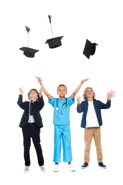 Niños vestidos con disfraces de diferentes profesiones lanzando gorras de graduación de aire aislados en blanco - foto de stock