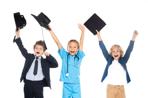 Crianças excitadas vestidas com trajes de diferentes profissões segurando gorros de graduação pretos acima das cabeças isoladas no branco — Fotografia de Stock