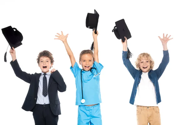 Crianças excitadas vestidas com trajes de diferentes profissões segurando bonés de graduação preto acima das cabeças isoladas no branco — Fotografia de Stock