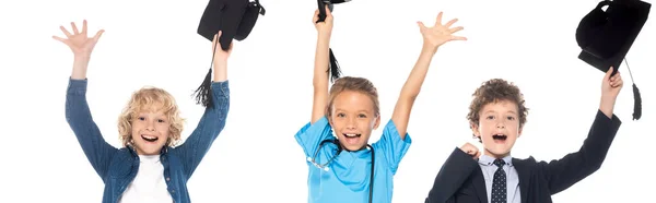 Plano panorámico de niños emocionados vestidos con disfraces de diferentes profesiones con gorras de graduación sobre cabezas aisladas en blanco — Stock Photo