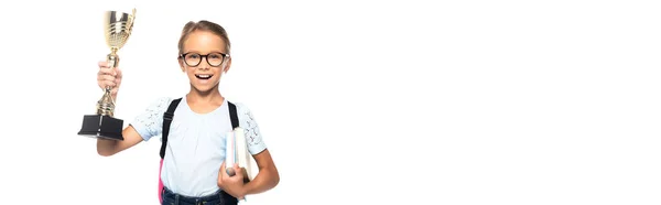 Panoramic concept of excited schoolgirl in glasses holding books and golden trophy isolated on white — Stock Photo
