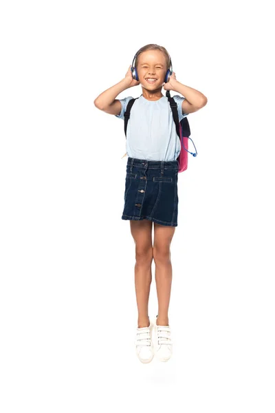 Colegiala con los ojos cerrados tocando auriculares inalámbricos mientras salta aislado en blanco - foto de stock