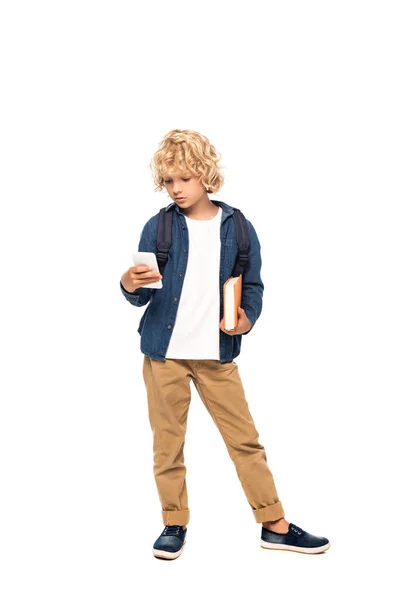 Blonde and curly schoolkid holding book while using smartphone isolated on white — Stock Photo