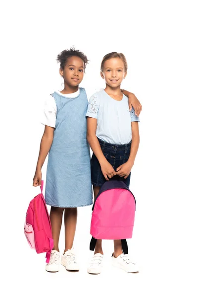 Multicultural schoolgirls holding backpacks while hugging isolated on white — Stock Photo