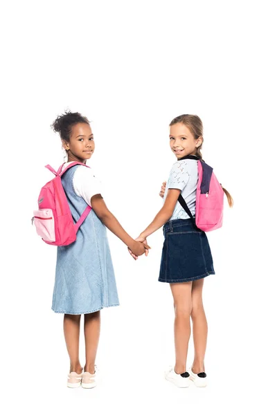 Escolares multiculturais com mochilas de mãos dadas e olhando para a câmera isolada em branco — Fotografia de Stock