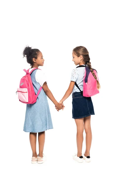 Multicultural schoolkids with backpacks holding hands and looking at each other isolated on white — Stock Photo