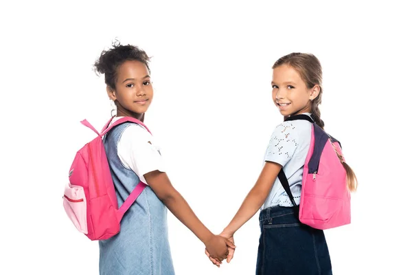 Amis multiculturels avec des sacs à dos tenant la main et regardant la caméra isolée sur blanc — Photo de stock