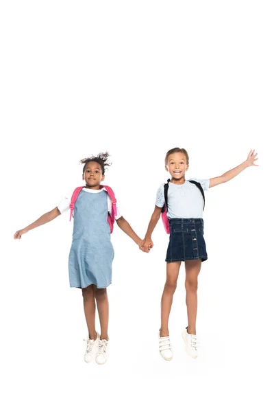 Colegialas multiculturales con mochilas cogidas de la mano y saltando aisladas en blanco - foto de stock