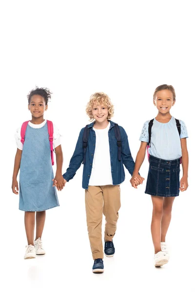 Blonde and curly schoolboy holding hands with multicultural schoolgirls while walking isolated on white — Stock Photo