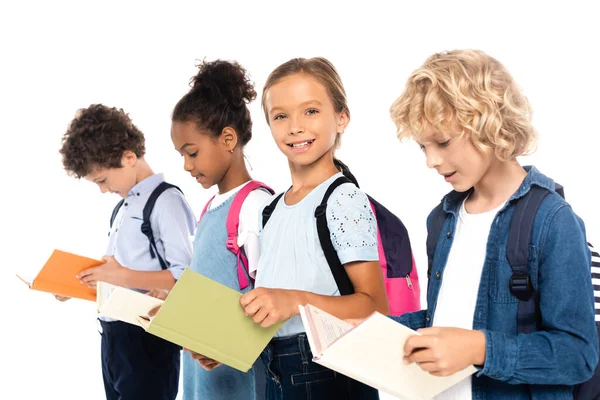 Enfoque selectivo de los escolares multiculturales con mochilas que sostienen libros aislados en blanco - foto de stock