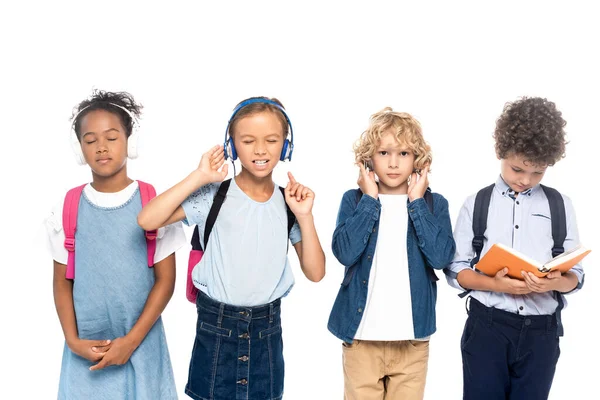 Multicultural schoolkids listening music in wireless headphones near curly boy reading book isolated on white — Stock Photo