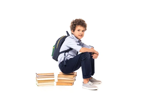 Lockiger Schuljunge mit Rucksack sitzt auf Büchern isoliert auf weißem Papier — Stockfoto